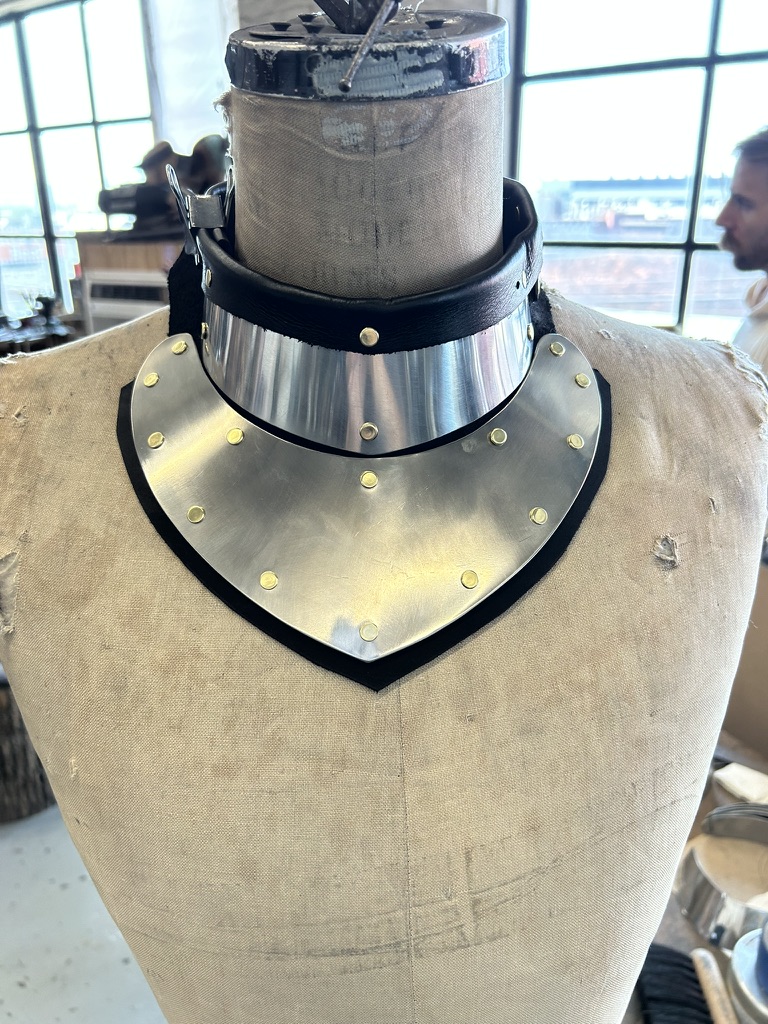 Polished steel gorget with brass rivets and black leather trim displayed on a mannequin