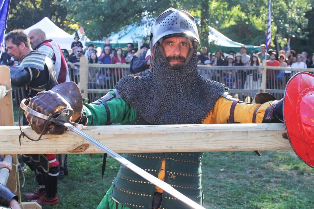 Sir Balin in medieval armor at an outdoor combat event, standing behind a wooden barrier with a red shield and weapon visible