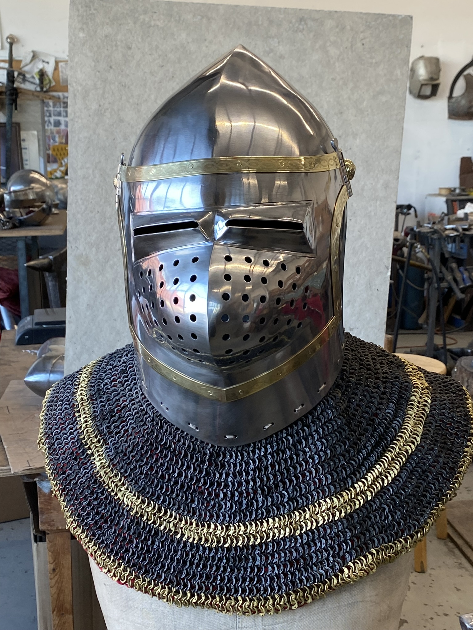Polished medieval helmet with brass trim displayed over dark chainmail in the workshop