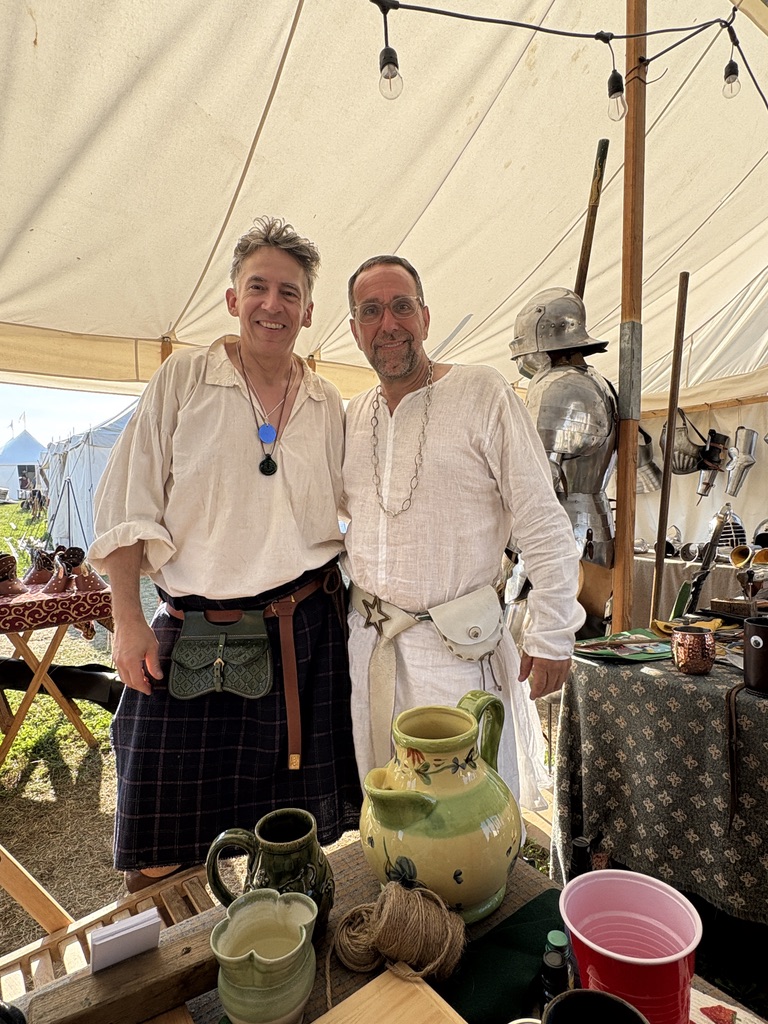 Master Jeffrey D. Wasson and a companion standing inside a medieval event tent with handcrafted armor displayed behind them