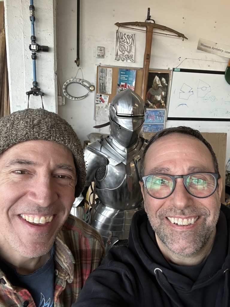 Two men smiling in a workshop with a polished suit of medieval armor behind them